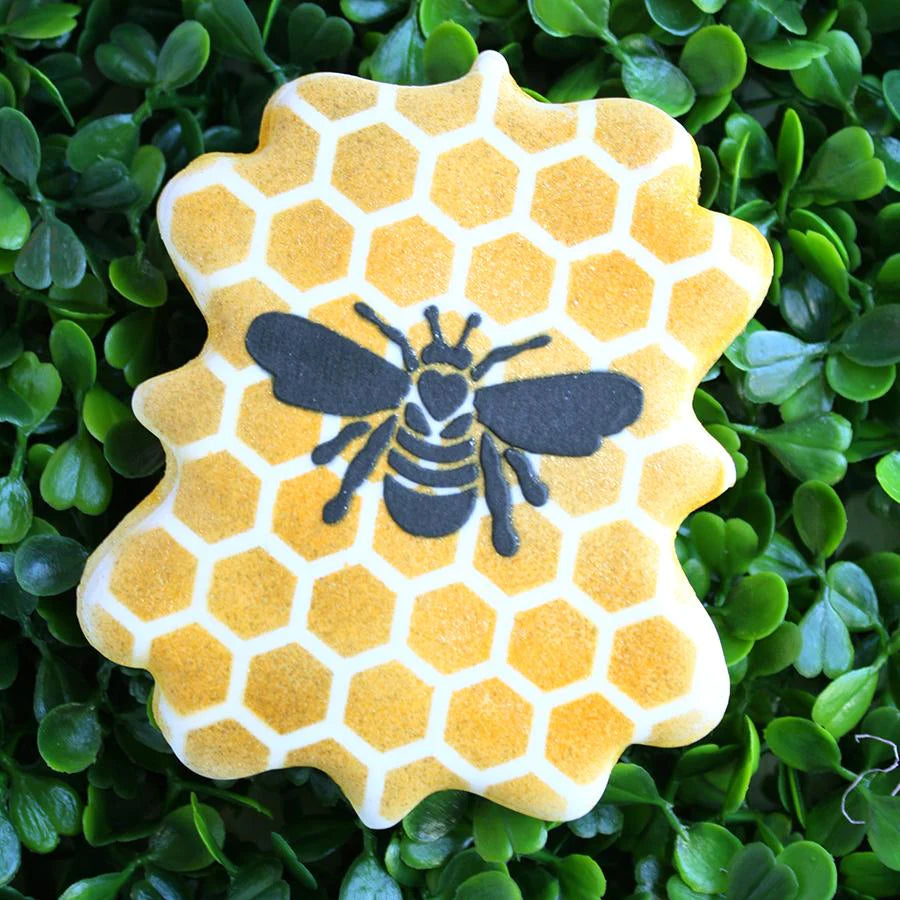 Sfeerbeeld van een prachtig gedecoreerd koekje met The Cookie Countess Bees stencil, honingraatpatroon in geel met een gedetailleerde bij in zwart en goud, gepresenteerd op groene blaadjes