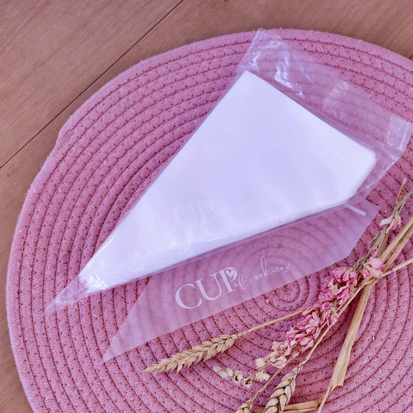 Verpakking tipless spuitzakken van Cup of Cookies met een losse spuitzak ervoor, gepresenteerd op een roze geweven onderzetter met gedroogde bloemen en tarwe-aren op een houten ondergrond.