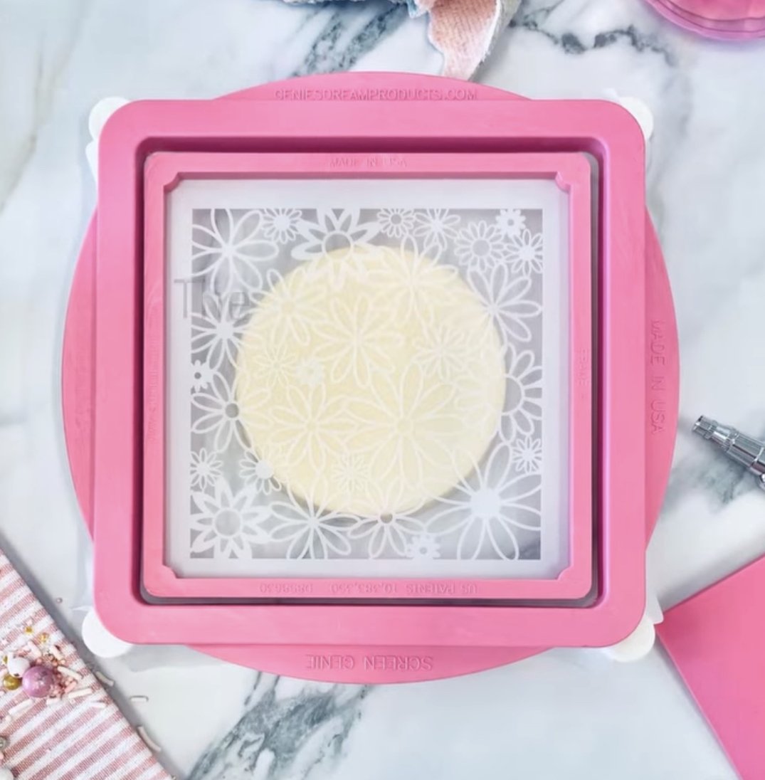 Roze Screen Genie van de Creative Cookier met fijn gaas over een bloemenstencil op een rond koekje