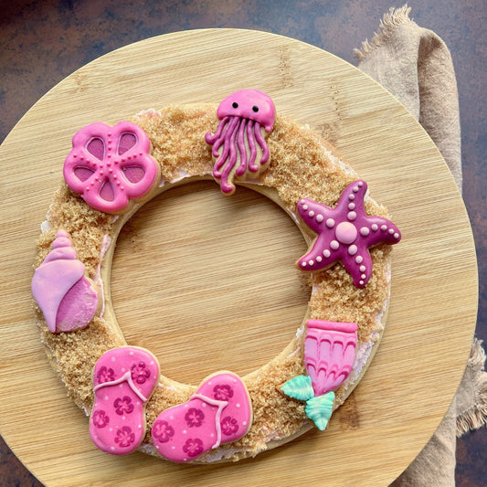 Koekkrans strand van Cup of Cookies met roze en paarse koekjes: wulk schelp, zeester, kwal, zand dollar, teenslippers en mermaid tail op een zandlaagje