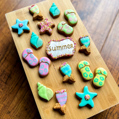 Mini zomerkoekjes van Cup of Cookies op een houten plank: wulk schelpen, zeesterren, mermaid tails, teenslippers en een Summer-bordje in turquoise, roze, geel en groen