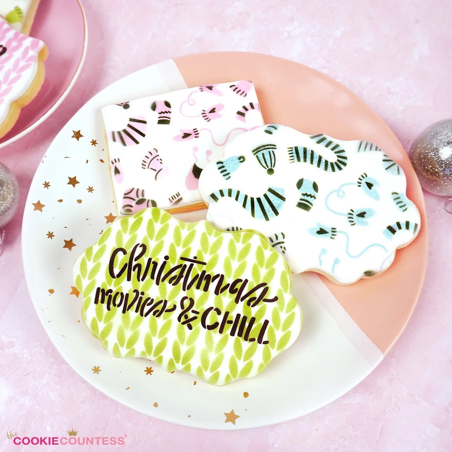 Sfeerbeeld van gedecoreerde kerstkoekjes met The Cookie Countess Christmas Movies and Chill stencil, kleurrijke koekjes met katten, wanten en kerstpatronen op een roze bord, perfect voor het decoreren van kerstkoekjes