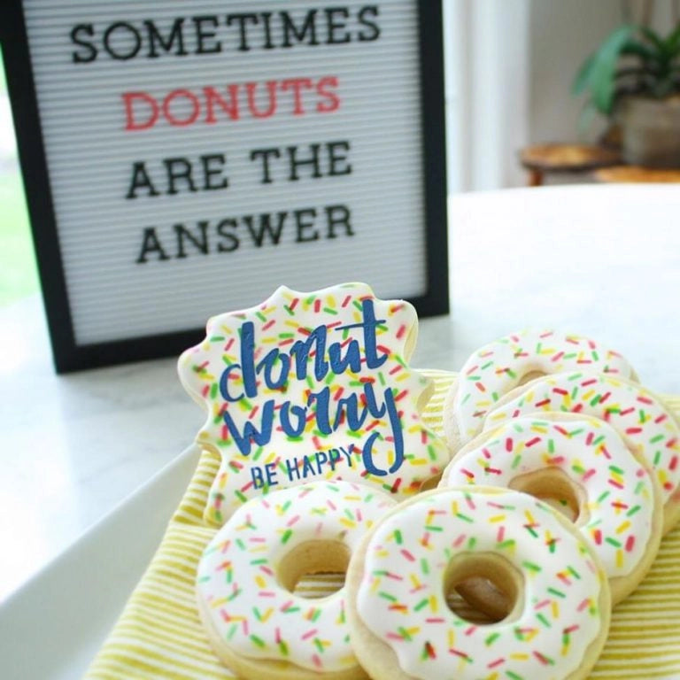 Sfeerbeeld van donutvormige koekjes gedecoreerd met The Cookie Countess Donut Worry Be Happy stencil, kleurrijke letters en hagelslag op witte donuts naast een letterbord met Sometimes Donuts Are The Answer