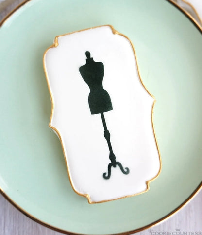 Sfeerbeeld van een elegant versierd koekje met The Cookie Countess Dress Mannequin stencil, zwart paspop silhouet op wit royal icing met gouden rand, gepresenteerd op een mintgroen bordje
