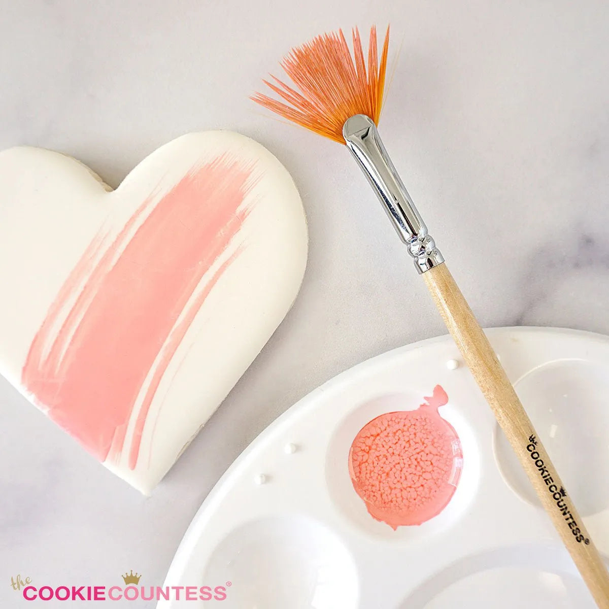Sfeerbeeld van The Cookie Countess Food Safe Fan Brush met houten handvat en oranje waaierborstelharen naast een gedecoreerd hartkoekje en verfpalet met roze eetbare verf, ideaal penseel voor het schilderen op koekjes