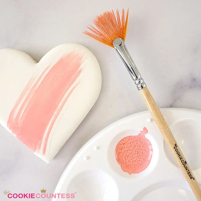 Sfeerbeeld van The Cookie Countess Food Safe Fan Brush met houten handvat en oranje waaierborstelharen naast een gedecoreerd hartkoekje en verfpalet met roze eetbare verf, ideaal penseel voor het schilderen op koekjes