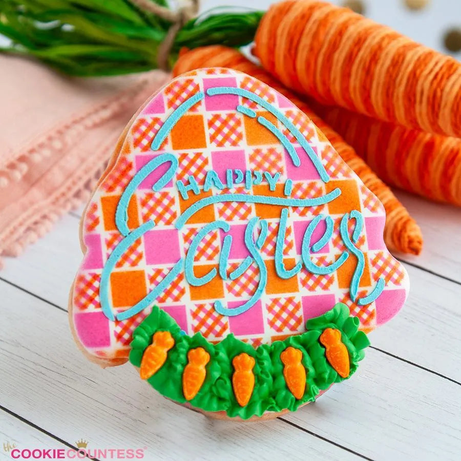 Sfeerbeeld van een paasei-vormig koekje gedecoreerd met The Cookie Countess Happy Easter PYO stencil, kleurrijk roze-oranje ruitpatroon met Happy Easter tekst in blauw royal icing en kleine worteltjes, gepresenteerd op een houten ondergrond