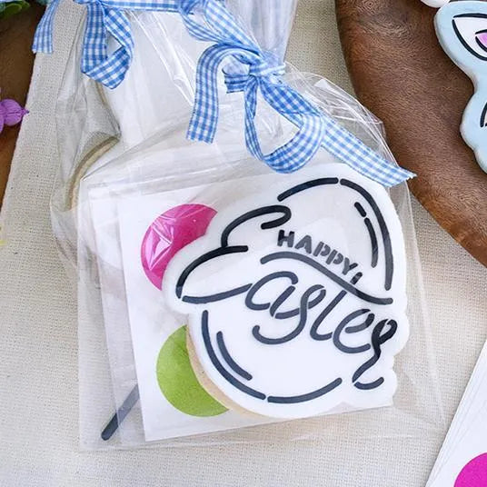 Sfeerbeeld van een koekje beschilderd met The Cookie Countess Happy Easter PYO (Paint Your Own) stencil, Happy Easter tekst in zwart op wit koekje verpakt in cellofaan zakje met blauw geruit lint, naast kleurrijke paaseitjes
