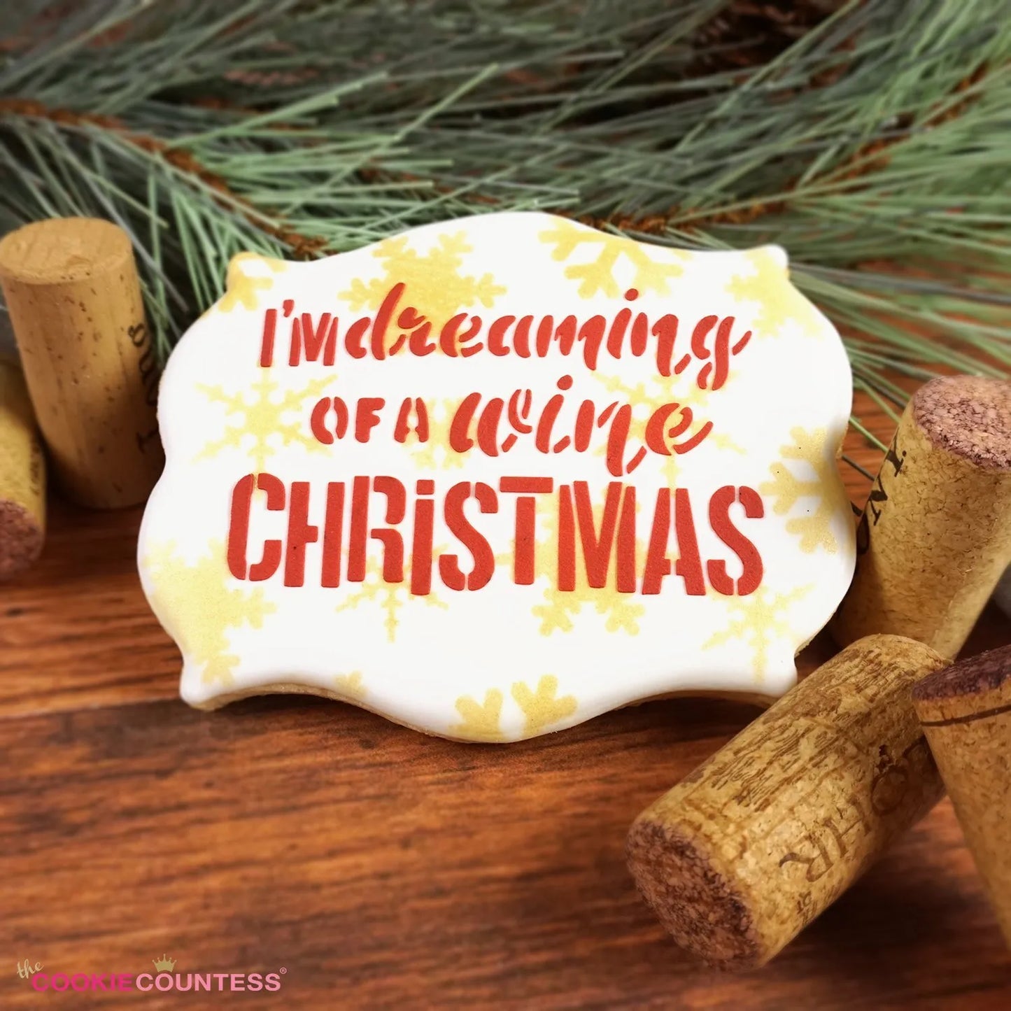 Sfeerbeeld van een gedecoreerd kerstkoekje met The Cookie Countess I'm Dreaming of a Wine Christmas stencil, sierlijk koekje met rode tekst en gouden sneeuwvlokken tussen wijnkurken en dennentakken, perfect kerststencil voor koekjes