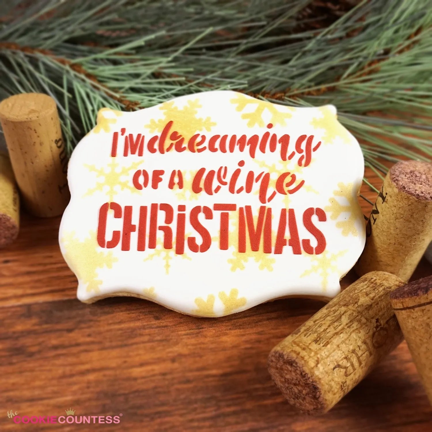 Sfeerbeeld van een gedecoreerd kerstkoekje met The Cookie Countess I'm Dreaming of a Wine Christmas stencil, sierlijk koekje met rode tekst en gouden sneeuwvlokken tussen wijnkurken en dennentakken, perfect kerststencil voor koekjes