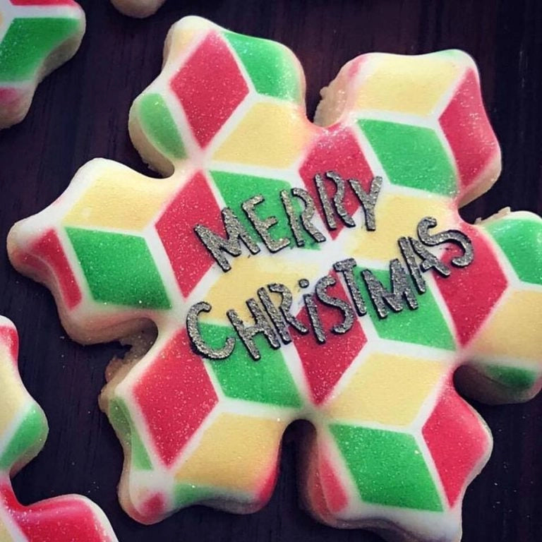 Sfeerbeeld van een sneeuwvlokvormig koekje gedecoreerd met The Cookie Countess Merry Christmas Funky stencil, kleurrijk ruitpatroon in rood, groen en geel met Merry Christmas tekst in zilveren glitterletters