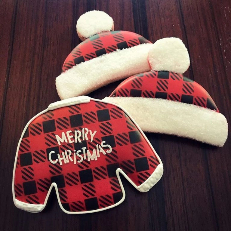 Sfeerbeeld van kerstmuts en kersttrui vormige koekjes gedecoreerd met The Cookie Countess Merry Christmas Funky stencil, rood-zwart buffalo plaid patroon met Merry Christmas tekst en witte royal icing details