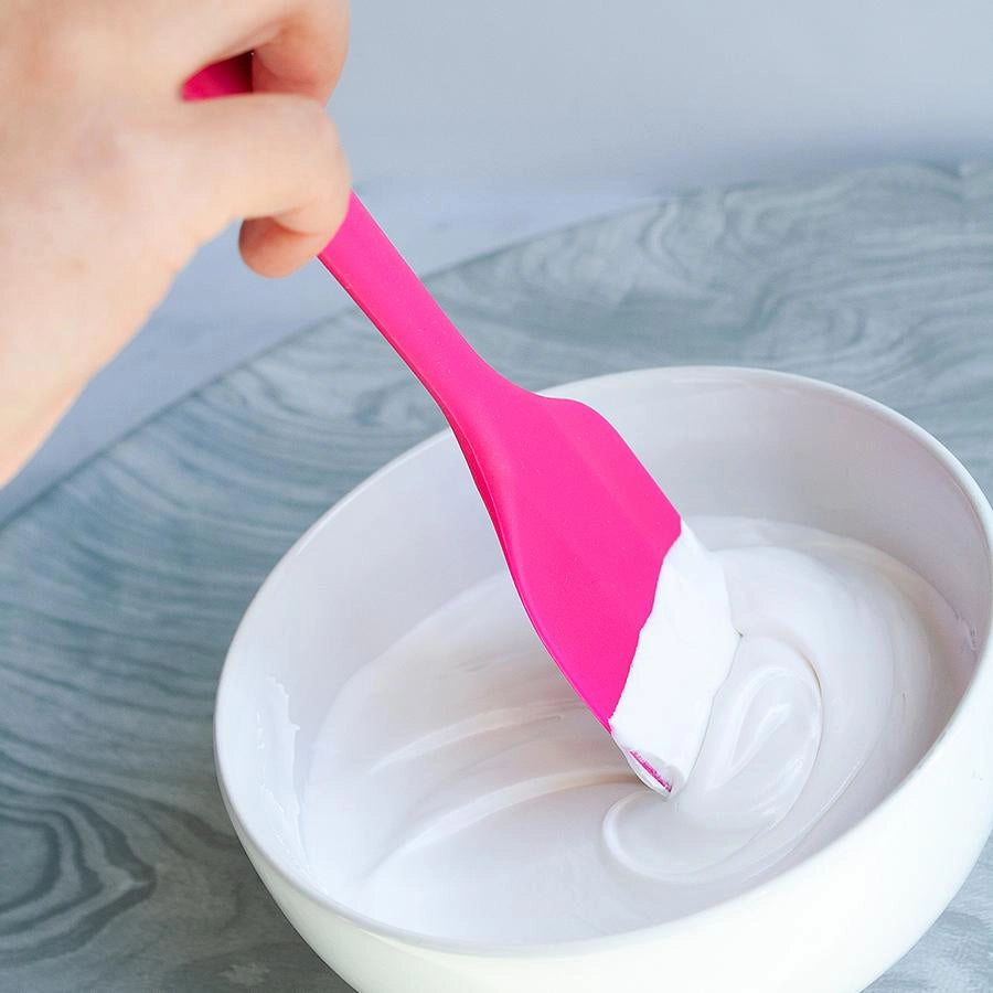 The Cookie Countess siliconen spatula in roze in gebruik, hand die de flexibele spatel gebruikt om witte royal icing uit een kom te scheppen, ideaal keukengerei voor het decoreren van koekjes