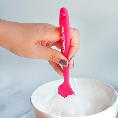 Sfeerbeeld van The Cookie Countess siliconen spatula in roze, hand die de spatula gebruikt om witte royal icing uit een kom te scheppen, ideale flexibele spatel voor het mengen en aanbrengen van royal icing op koekjes