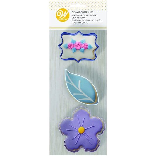 Wilton Cookie Cutter Set Floral verpakking met drie bloemen-thema koekjesuitstekers en versierde voorbeeldkoekjes: een sierlijk frame met roze bloemen, een blauw blad en een paarse bloem, metalen uitstekers voor lente- en zomerkoekjes
