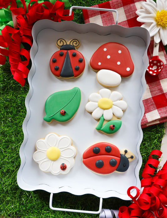 Zes Love Bug koekjes met royal icing op een wit dienblad: lieveheersbeestjes, madeliefjes, paddenstoel en blaadje