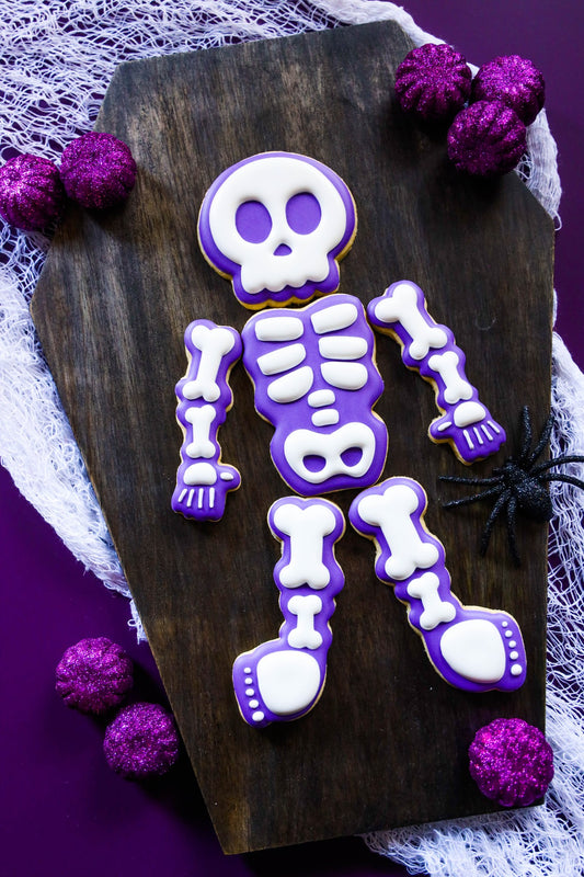 Halloween-skelet van zes koekjes met paarse en witte royal icing: schedel, romp, armen en benen op een houten plank